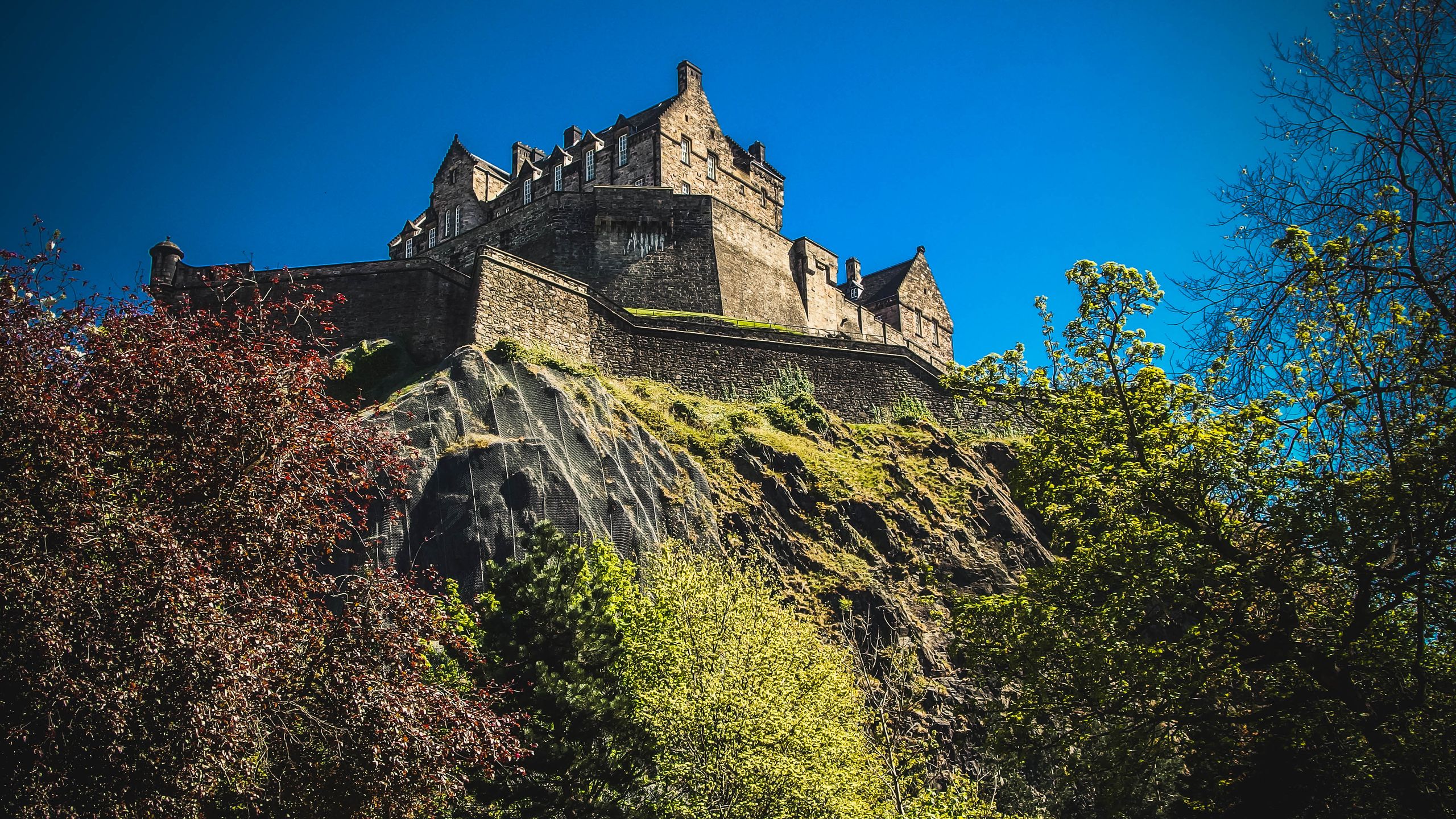 Iconic Edinburgh Castle atop Castle Rock under a clear blue sky, surrounded by lush greenery.
